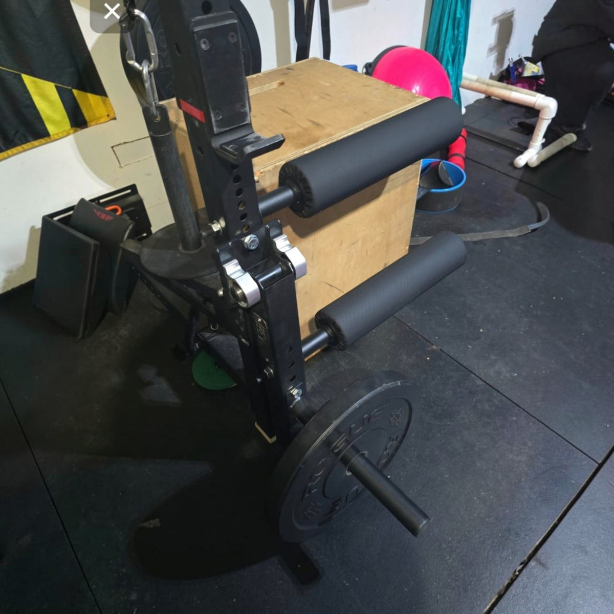 Gymless black steel leg curl and extension attachment with foam rollers mounted on a gym rack with a weight plate.