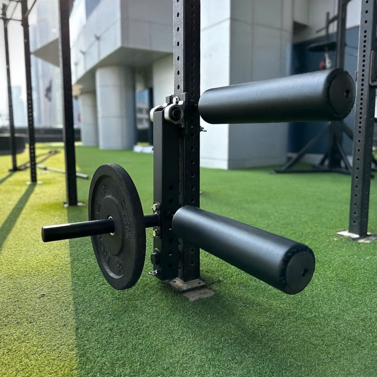 Gymless leg curl and extension attachment mounted on a black power rack with weight plate on an outdoor gym turf
